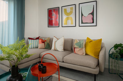 Foxes Chase showhome living room with light grey L-shaped sofa, multicoloured abstract cushions, three framed abstract prints above, teal sheer curtains on the left, and nested red and green circular coffee tables on a patterned rug.