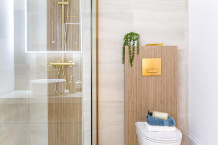 Walk-in shower with frameless glass screen, gold thermostatic shower valve and handset on wood-effect wall tile; adjoining concealed-cistern WC with matching wood panelling and gold dual-flush plate, topped with a cascading succulent, folded white and teal towels, and a luxury hand-wash bottle.