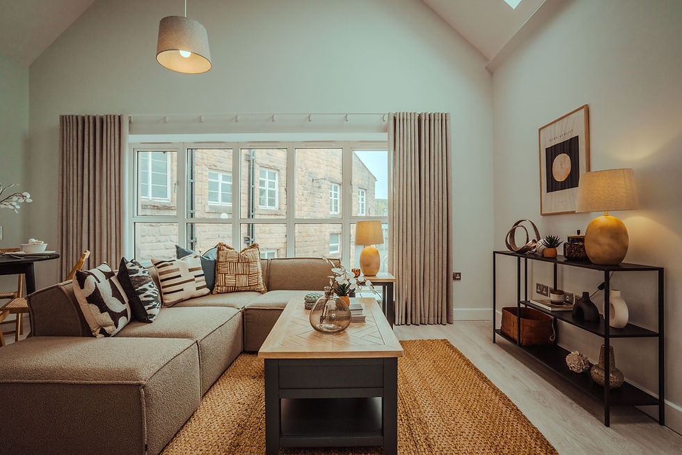 Cozy living room with beige sectional sofa, patterned cushions, wooden coffee table, large window, woven rug, and warm lighting.