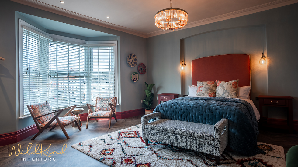 Cozy bedroom with a red headboard, patterned chairs, and a chandelier. Large bay window on the side. "WildKind Interiors" logo visible.