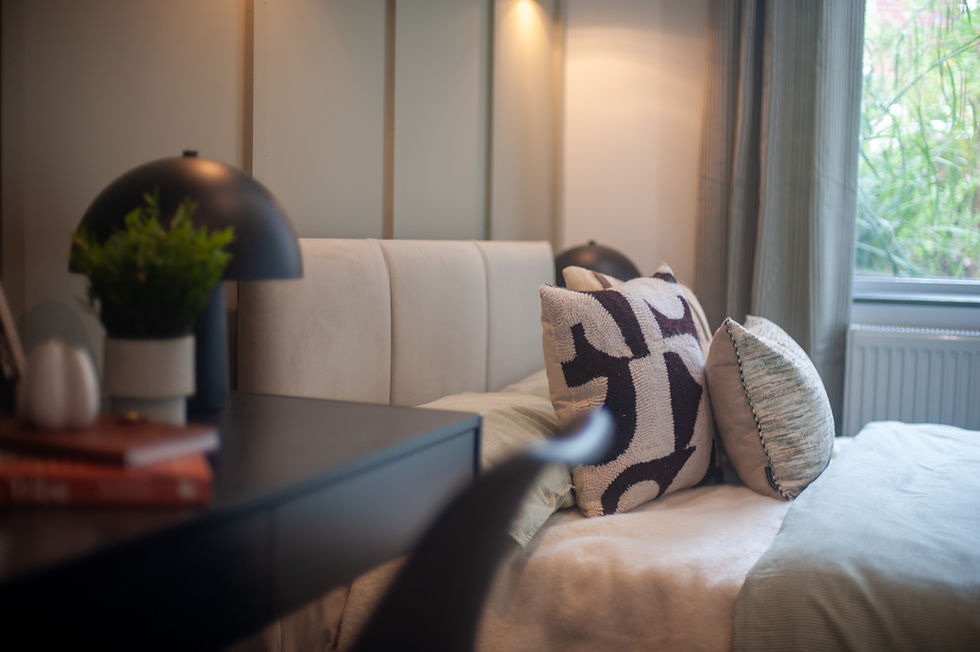 Cozy bedroom with a bed adorned with patterned pillows, a sleek lamp, and books on a side table. Soft lighting and window view.