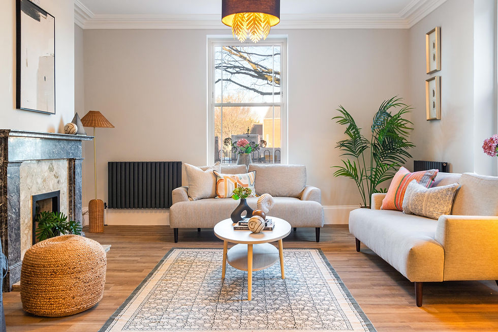 Cozy living room with beige sofas, colorful cushions, a patterned rug, and a round wooden table. Large window, plants, art on walls.