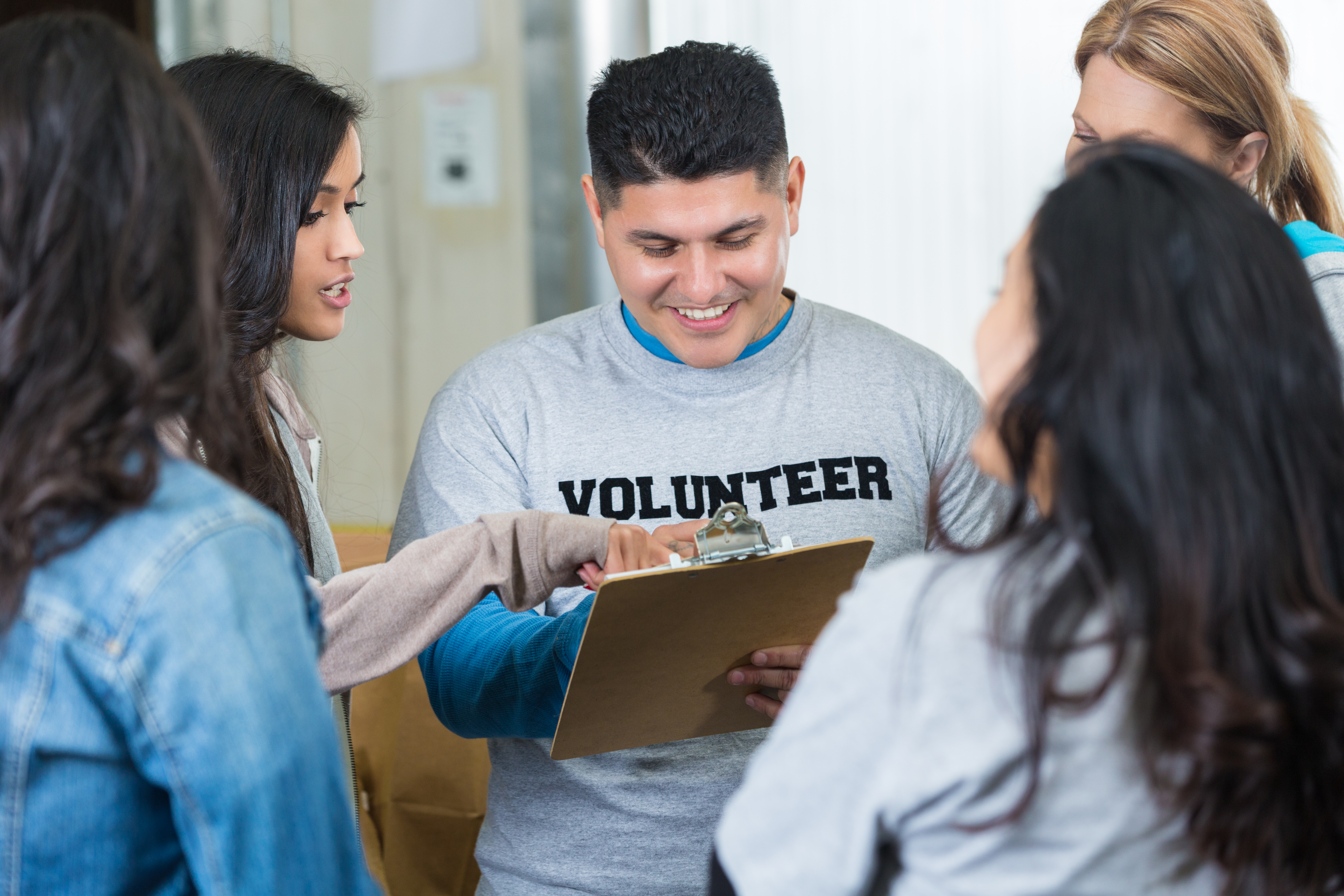 A man with a clipboard and shirt reading "volunteer" leads a group meeting with other volunteers.