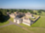 Drone photo of a castle gatehouse sitting before a Tudor country house surrounded by castle walls and set upon a backdrop of green hills and countryside
