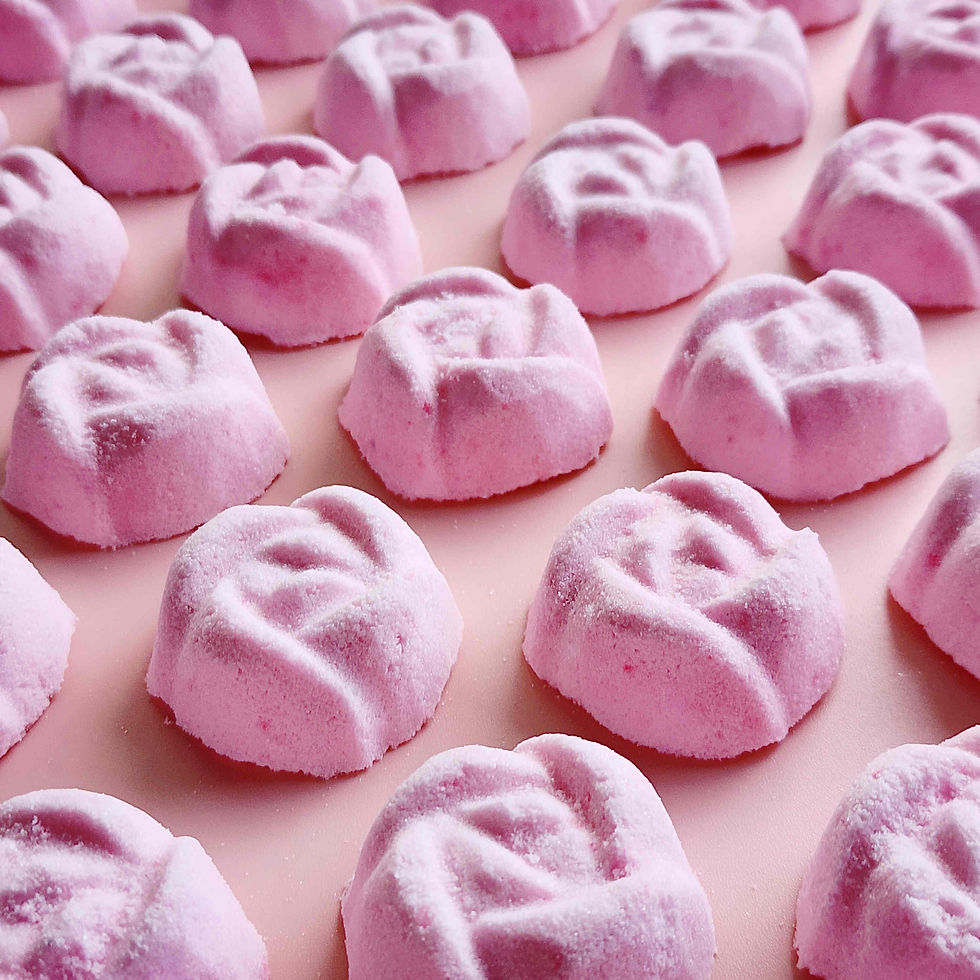 Thumbnail: light dusty pink coloured mini bath bombs in the shape of roses laid out neatly on a plain pink background