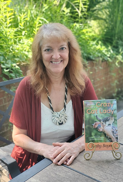 Kathy is sitting at a table outside with her book, Memoir of a Crazy Cat Lady, upright in a stand on the table in front of her.