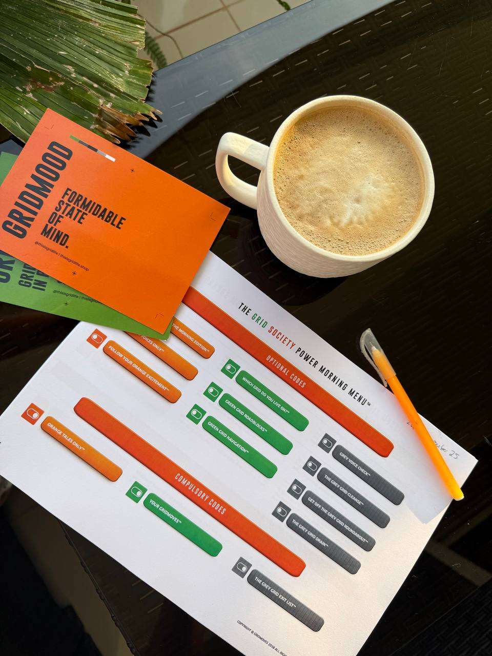 Coffee cup beside Grid Society workbook pages and an orange notebook on a dark table, representing quiet confidence, self-development, and no longer feeling ashamed of your current position while building your future behind the scenes.