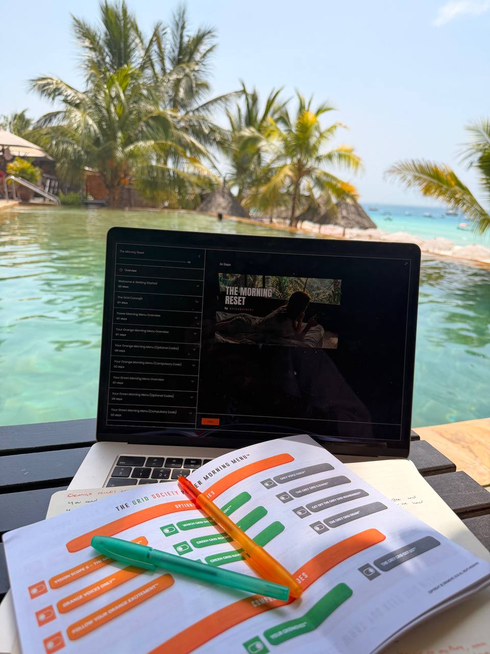 Laptop and Grid Society Morning Reset workbook with orange and green pens beside water and palm trees, representing structured morning reflection.