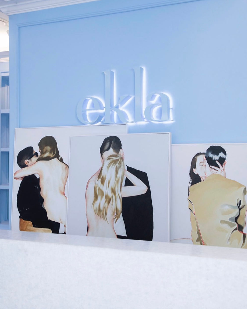 Three paintings of couples kissing are displayed against a light blue wall. Neon text "ekla" is above, creating a chic and romantic atmosphere.