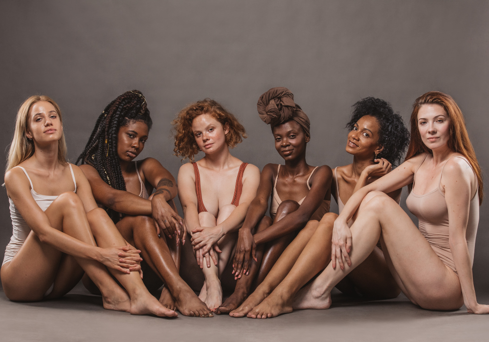 A group of women from different ethnic backgrounds sit together against a gray background. The women are wearing neutral-toned leotards and look relaxed.