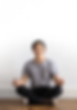 A man sits cross-legged on the ground in a yoga pose with his hands resting on either knee. He wears a gray tee shirt, dark jeans, and gray sneakers. He sits on a hardwood floor against a white wall.