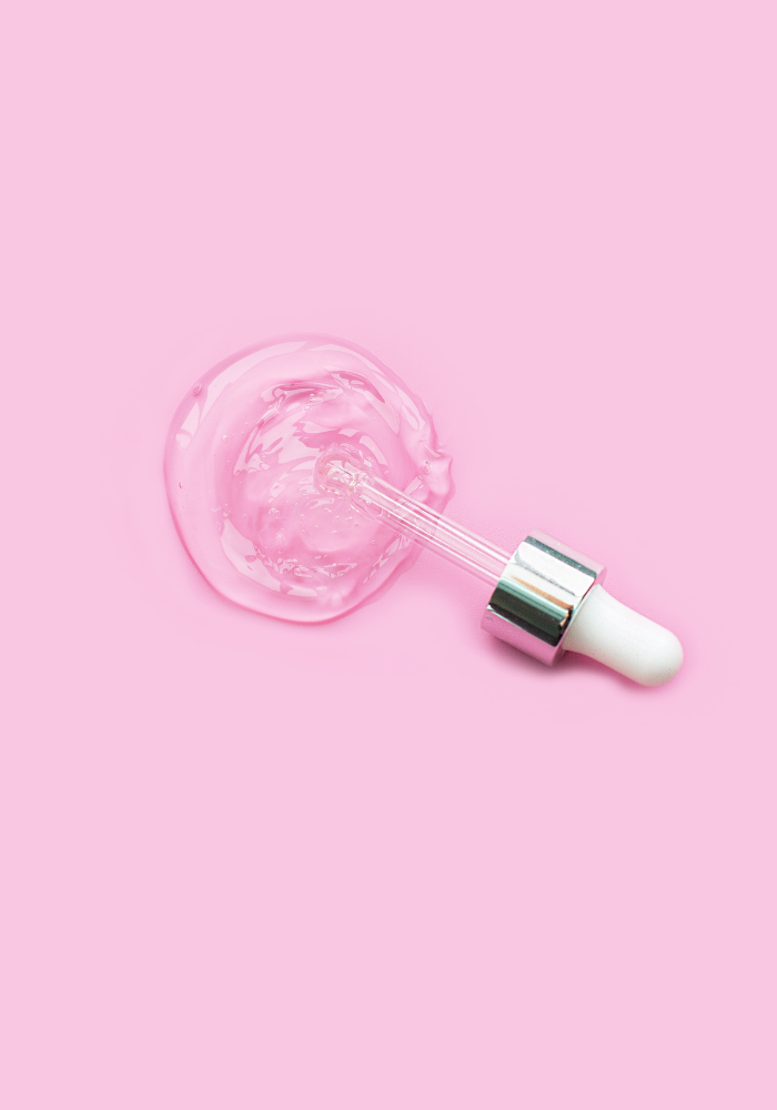 A white and silver dropper expels clear gel in a puddle on a pink backdrop.