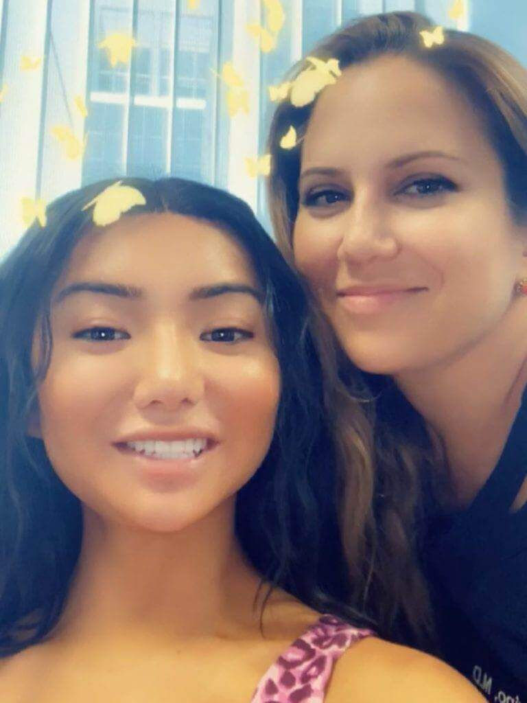 Dr. Sarah Yovino poses with Nikita Dragun in a medical office. A filter is applied to the photo, adding glowing golden butterflies around each woman's head.