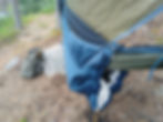 Eine Hängematte mit einem Underquilt ist im aufgespannten Zustand zu sehen. Mit im Bild ist ein Rucksack und das noch nicht aufgespannte Tarp. Im Hintergrund sieht man das Ufer eines Waldsees mit Seerosen.