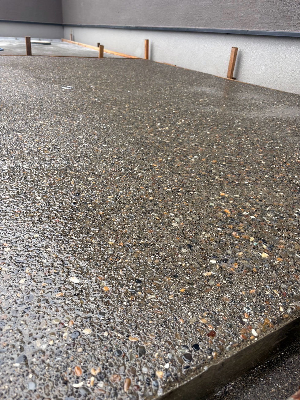 雨に濡れた洗い出し仕上げのコンクリート床が手前に広がり、奥に灰色の基礎と木の枠が並ぶ工事中の場面