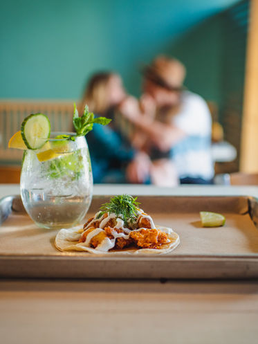 Product photography of a taco and a drink with couple feeding eachother tacos in the background.
