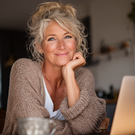 Smiling woman with gray hair, wearing a cozy sweater, leans on hand by a laptop in a warmly lit room, conveying a relaxed mood.