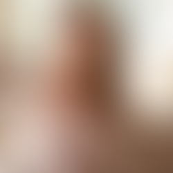 Woman with long hair smiling at a laptop in a bright room, wearing a beige sweater and gold necklaces. Flowers and a mug are nearby.