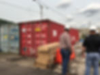 Two people in hardhats standing next to 5 stacked boxes and 2 shipping containers