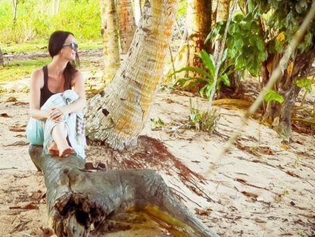 Sarah Harris sitting on a fallen tree beneath palm trees on a tropical beach in Indonesia.
