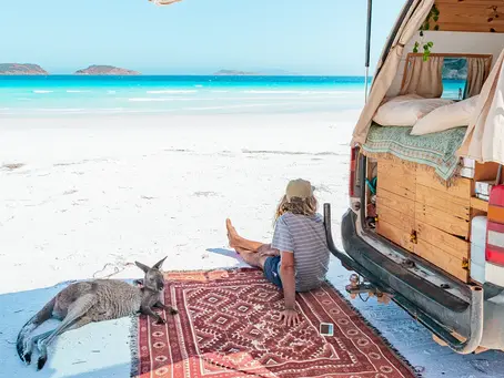 Woman relaxing beside a campervan and a kangaroo on a white sandy beach overlooking turquoise water in Western Australia.