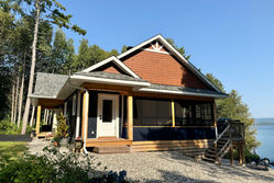 Exterior elevation of a one storey house overlooking the water. Brown/ reddish wood siding with a gable detail. Light grey shingle and a covered front entry. 