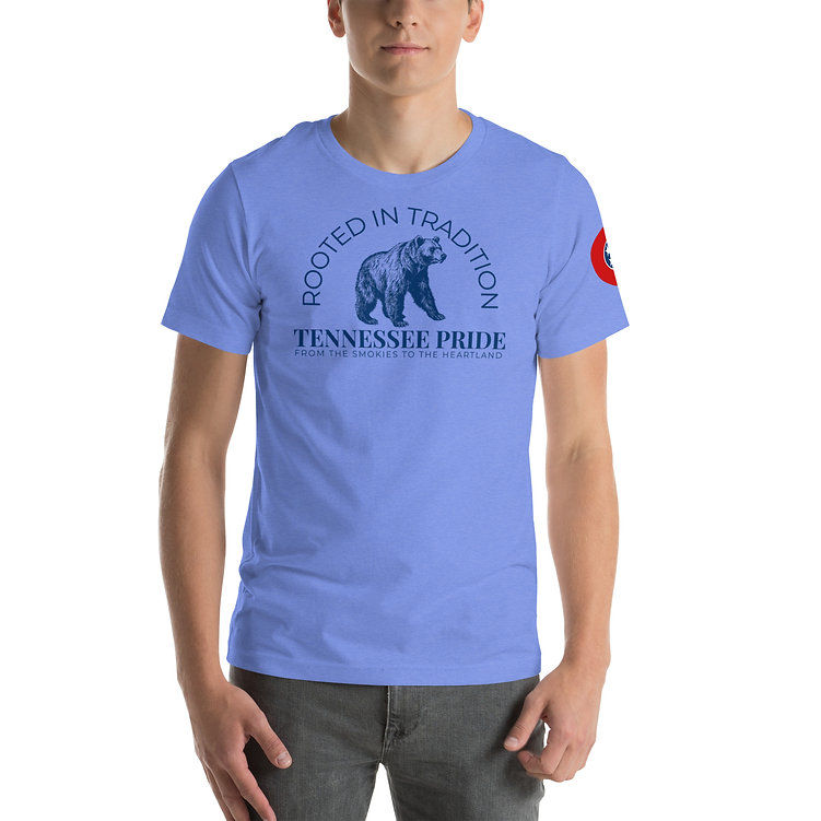 A man wearing a light blue T-shirt that reads “Rooted in Tradition” above an illustration of a bear. Below the bear, the text says “Tennessee Pride – From the Smokies to the Heartland.” The shirt also features a red Tennessee state flag emblem on the sleeve.
