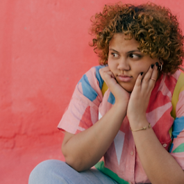 Woman wearing a colourful top sitting in front of bright red wall with head in hands, considering