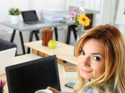 I woman with blonde hair and brown eyes sitting at a desk and looking over her shoulder with a slight, ironic smile on her face while she holds a mug of tea.