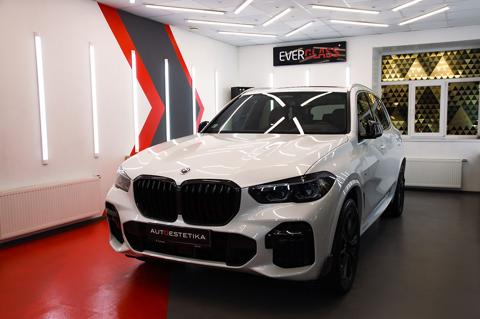 Baltas auto stāv telpā ar modernām sarkanām un pelēkām sienām. Redzams "EVERGLASS" un "AUTOESTETIKA" teksts. Spilgtas gaismas.