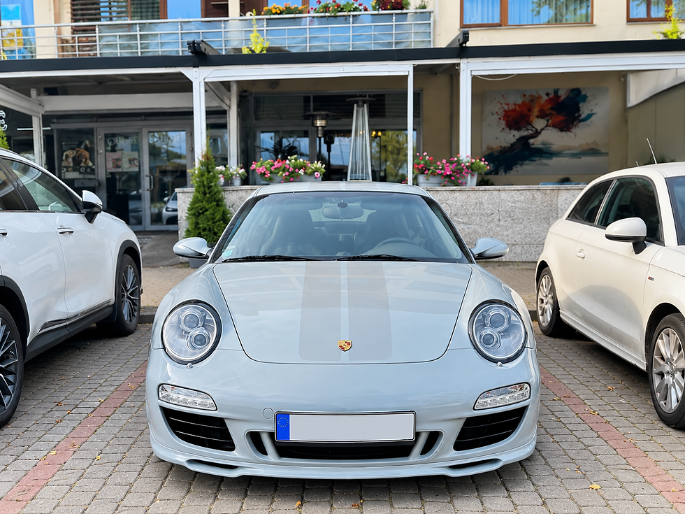 Sudraba Porsche stāv starp diviem baltiem auto pie viesnīcas ieejas ar ziediem un Eiropas numura zīmi.