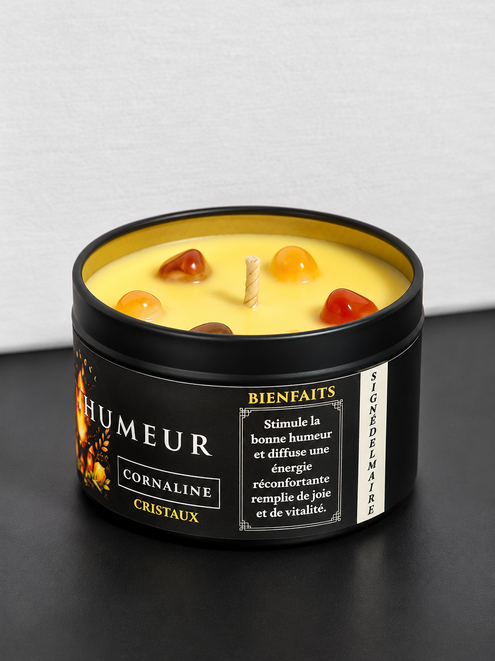 Bougie Bonne humeur à la cornaline et au parfum de vanille, dans un pot noir posé sur un livre, avec son couvercle à côté.
