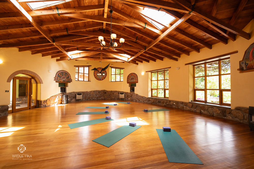 A yoga shala at Willka T'ika: a vaulted timber ceiling with skylights, warm plaster walls hung with carved pieces, tall windows onto the garden and mats laid out on a polished wooden floor