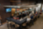 Bar setting with people seated, staff behind the counter. Blue lights, TV showing sports, text reads "WINE COFFEE BEER". Relaxed vibe.