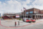 Brick building with "Stage 12" sign, flags, and restaurant vans in parking lot. Overcast sky, modern urban setting.