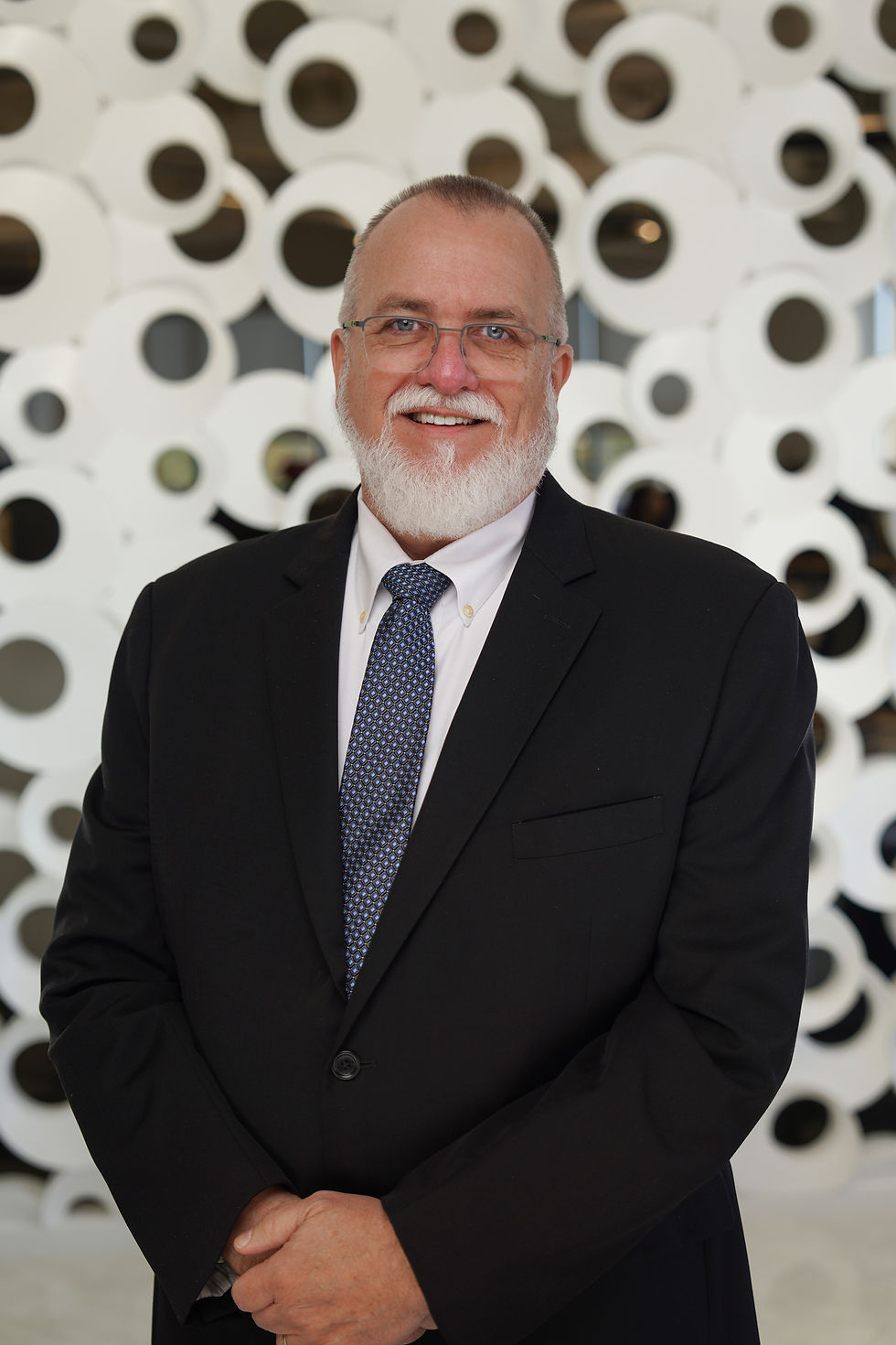 Man in a suit stands smiling with hands clasped. Background features a modern wall of white, circular patterns. Mood is professional and friendly.