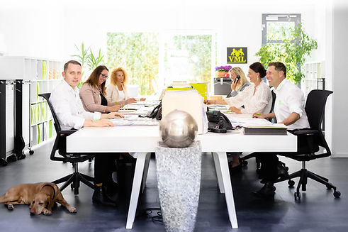 Die Mitarbeitenden und die Geschäftführer sitzen gemeinsam an einem großen, weißen Tisch. Dort sind mehrere Arbeitsplätze und Telefone und unterlagen sind zu sehen. Im Vordergrund steht eine dekorative Säule. Links vorne liegt ein brauner, schlafender Hund.ein brau