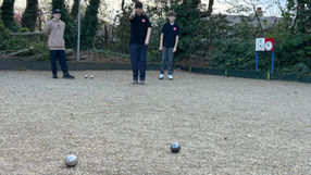 Three juniors play pétanque on a gravel court. One is throwing a silver boule. Trees and a scoreboard are in the background.