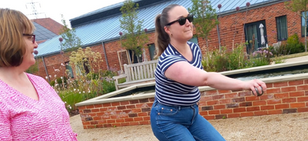 Two women, one wearing sunglasses and striped shirt, throwing a boule. Brick building and plants in the background.