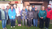 A group of 12 ladies stand together outdoors in front of a shed. Some are holding trophies. They are dressed in colorful jackets, smiling in sunlight.