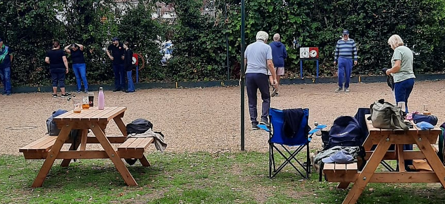People play pétanque on a gravel piste , with picnic tables, folding chairs, bags, and a leafy hedge behind them.