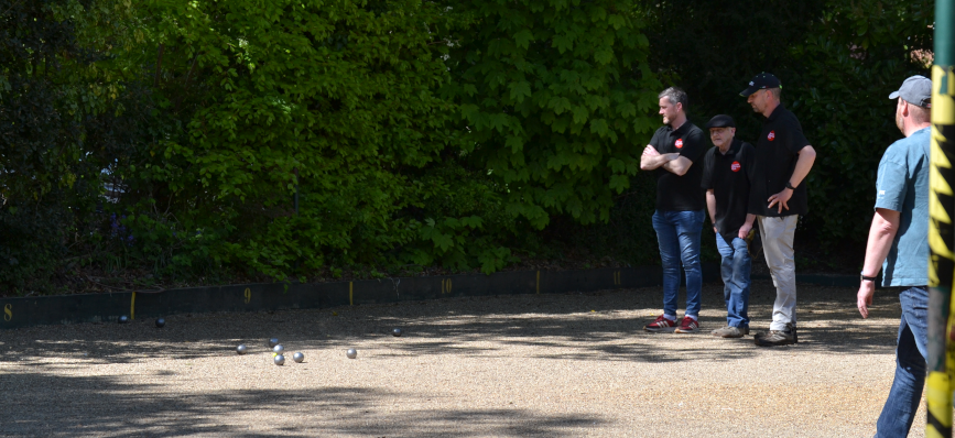 Four people playing pétanque, focusing on the boules scattered on a gravel court. Green foliage in the background. Bright, sunny day.