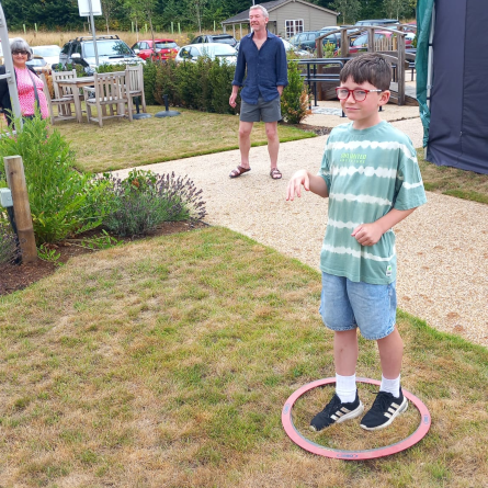 Boy in striped shirt stands in a ring on grass, hands raised. Adults watch in a garden setting with cars and buildings in the background.