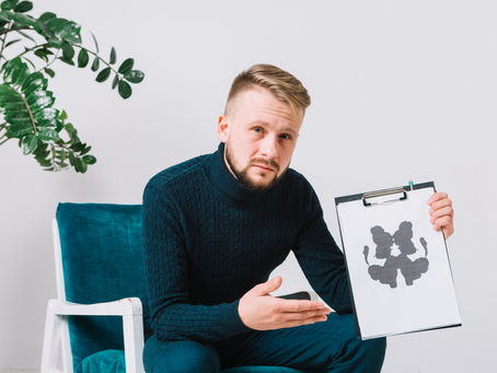 Man in blue sweater holds Rorschach inkblot on clipboard, appearing thoughtful. He sits in a blue chair, with a green plant in the background.