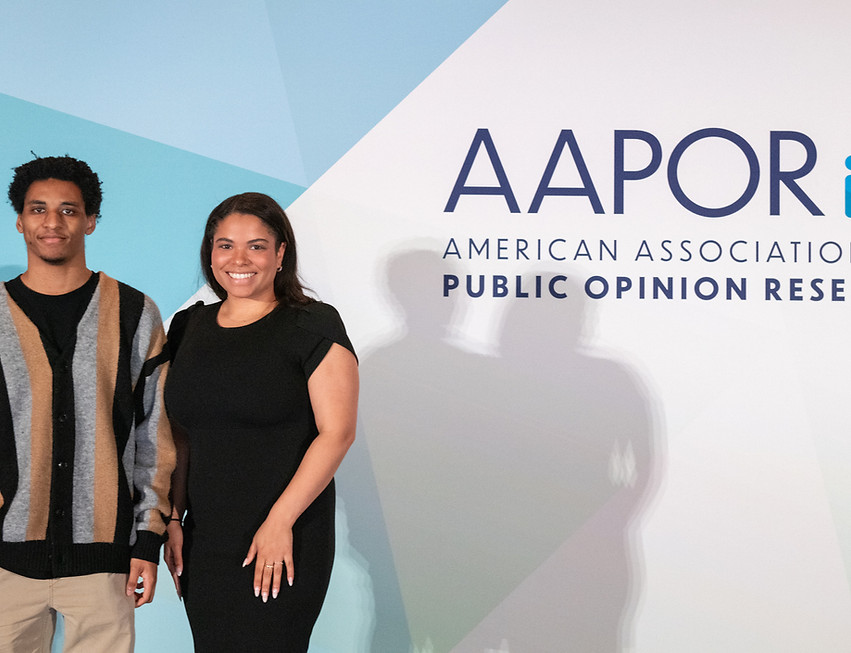Two people stand in front of AAPOR American Association for Public Opinion Research banner.