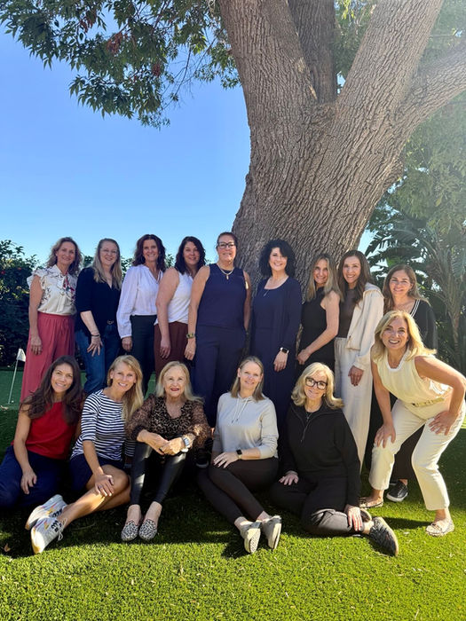 Group of diverse women posing together outdoors on green grass by a tree.
