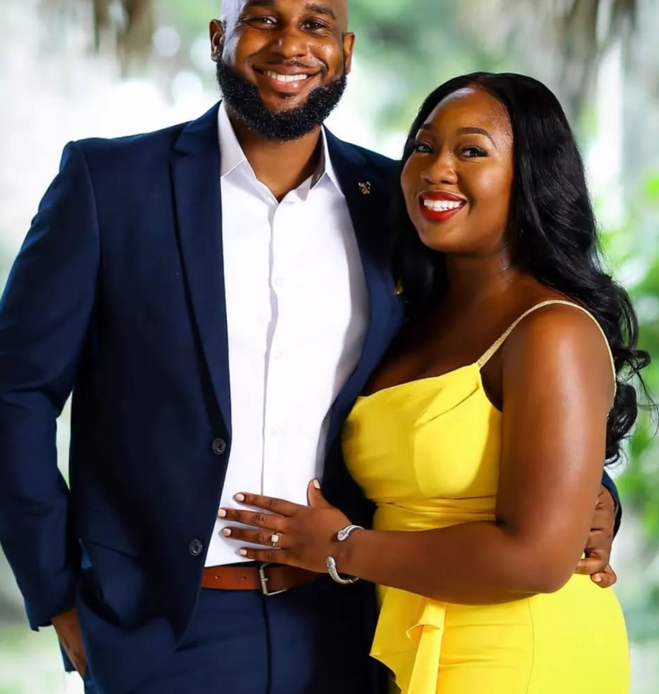 Smiling couple: man in suit, woman in yellow dress. Background with greenery.
