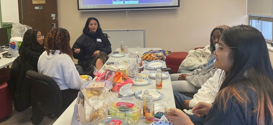 Group of students chatting and eating around a table with snacks.