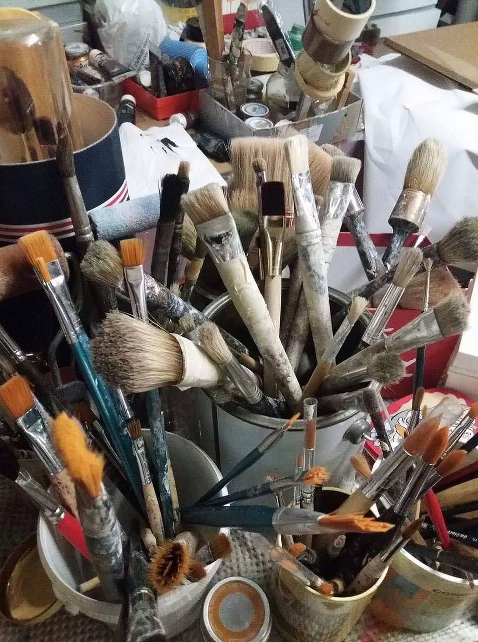 Contenitori pieni di pennelli di varie dimensioni e forme con setole usurate e manici dipinti, disposti in un ambiente artistico caotico.