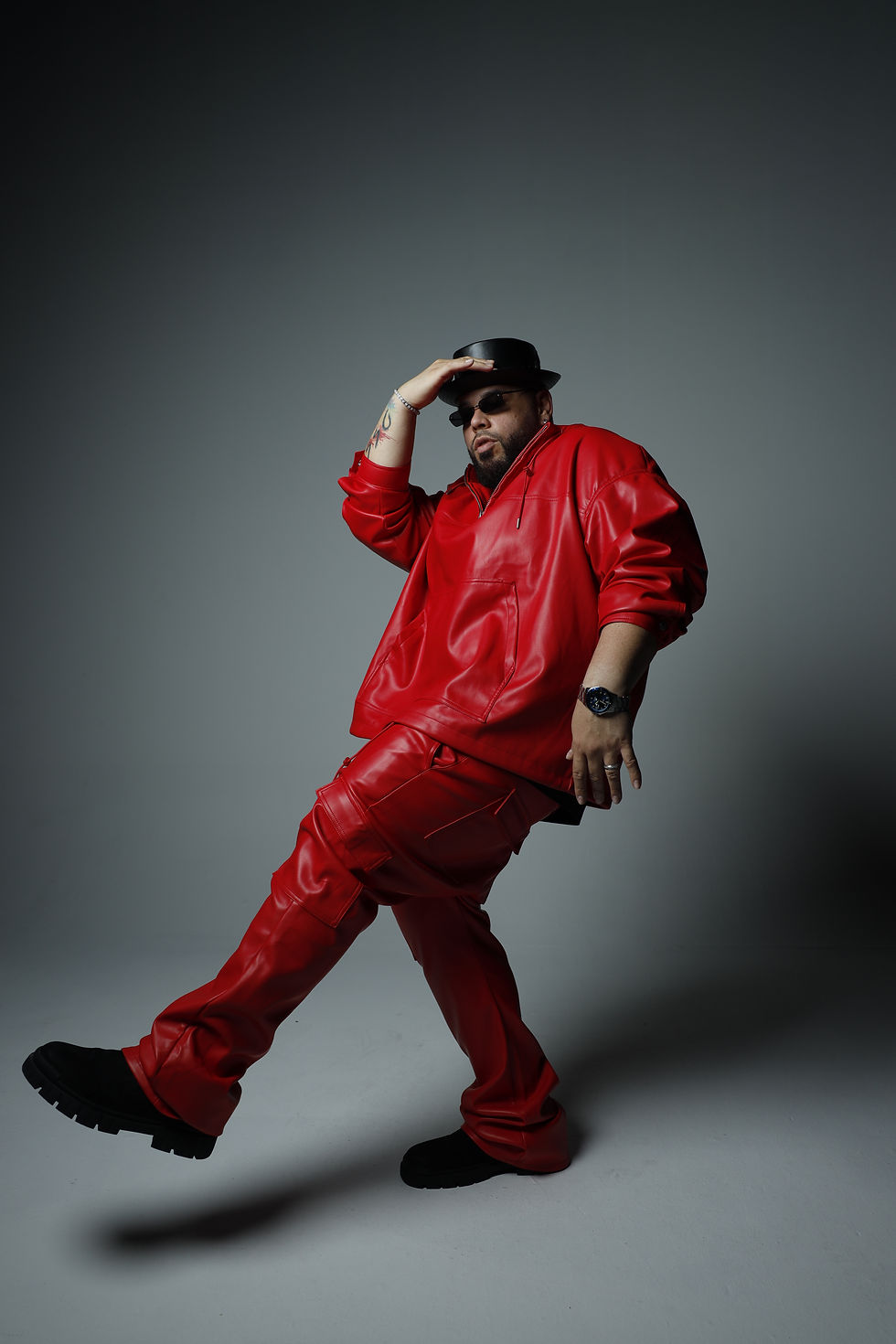 Pinto Picasso stands in a red leather jacket and pants leaning back with a black hat and sunlasses